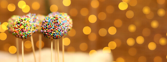 Colorful cake pops with bright sprinkles are displayed on sticks, adding joy to a festive holiday gathering. The warm lights create a cheerful atmosphere for Christmas and New Year 2026