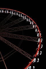 Diagonal view of a brightly lit Ferris wheel at night. The red and white lights highlight the structure against the dark sky, capturing the festive and modern city atmosphere.