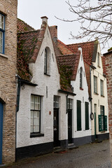 Charming row of whitewashed historic houses with red tiled roofs and green shutters on a narrow cobblestone street in Bruges, Belgium. The scene captures the peaceful atmosphere 