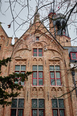 Gothic-style brick facade of a historic building in Bruges, Belgium, featuring arched windows with colorful stained glass and architectural details typical of the Flemish medieval heritage. 