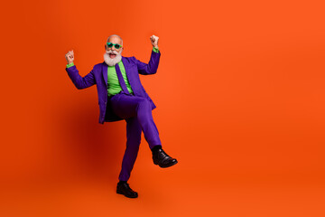 Elderly man in a purple suit dancing with joy against a bright orange background wearing a green...