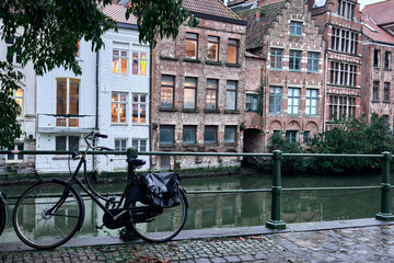 A scenic view of traditional Flemish buildings along the River Lys in Ghent, Belgium. A classic bicycle is parked by a green railing, surrounded by cobblestone and calm waters, under leafy branches. 