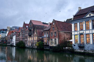 A row of historic houses lines the River Lys in Ghent, Belgium, captured during dusk. Their warm interior lights contrast with the cool evening sky