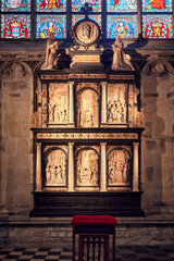 Detailed view of a stone altarpiece inside Notre-Dame du Sablon Church in Brussels, Belgium. The altarpiece features carved biblical scenes framed by columns, with colorful stained glass windows