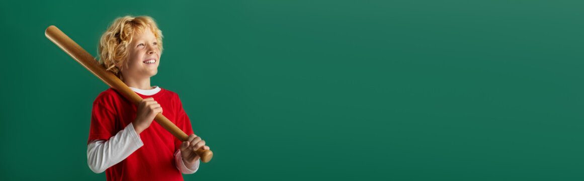 Joyful blonde boy in red shirt holding a wooden bat against a green background