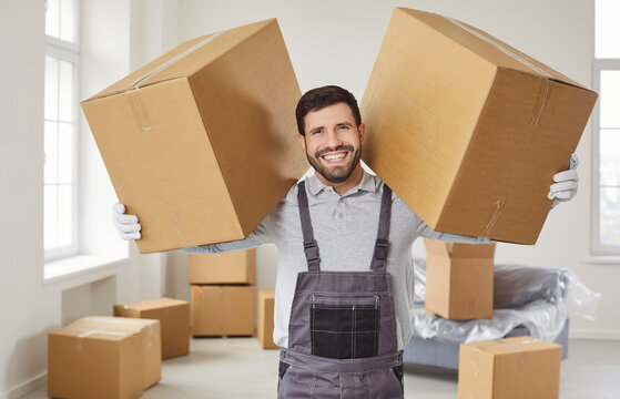 Happy man from moving company holding big cardboard boxes, standing inside of house, helping clients with removal or relocation, wearing uniform, unloading packages. Delivery and moving day 