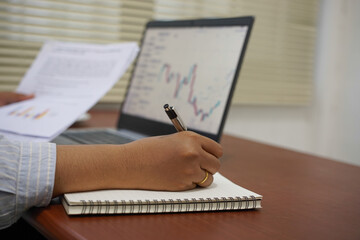 Selective focus on a woman’s hand writing notes near a white coffee mug with a blurry laptop displaying financial charts on a home office desk, concept for study and trading