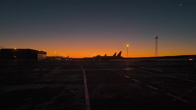 Winter airfield at dawn: dark apron, lights of masts and silhouettes of airplanes against the background of a bright orange strip of sky and a thin moon.
