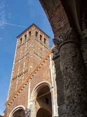 A low-angle shot captures the medieval red brick bell tower of the Basilica of Sant'Ambrogio in Milan, framed by a stone arch in the foreground.