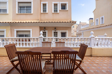 Outdoor tiled terrace with wooden dining furniture and white balustrade near beige residential buildings