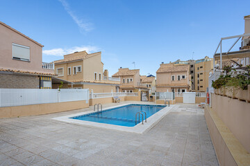 Private residential swimming pool with beige houses and tiled terrace under clear blue sky