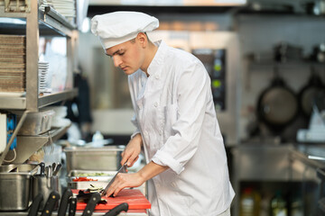 Guy cook in kitchen finely slices food and prepares ingredients for cooking. Kitchen worker in white uniform performs tasks of chef and auxiliary functions