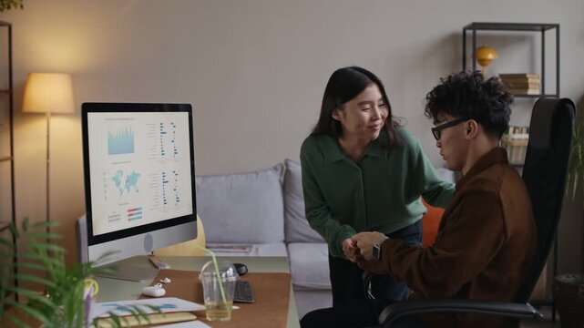 Medium long shot of young Asian man working on computer with financial screen in office and exchanging red envelopes with female colleagues during Tet celebration in startup office
