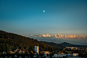 Suburban cityscape below the majestic, snow-capped Swiss Alps, illuminated by the setting sun. The half-moon hangs in the clear blue sky, blending urban life and nature.