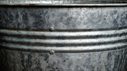 Close-up of a weathered, galvanized metal bucket, showing its textured surface, horizontal ridges, and visible rivets. The industrial aesthetic is prominent.