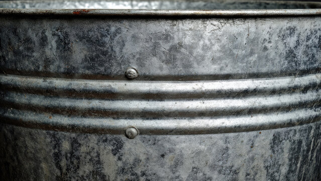 Close-up of a weathered, galvanized metal bucket, showing its textured surface, horizontal ridges, and visible rivets. The industrial aesthetic is prominent. - Powered by Adobe