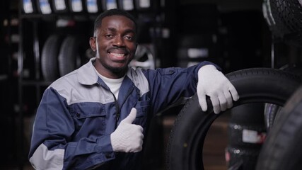 Happy African male adult mechanic with tire in auto shop giving thumbs up in tyre store.