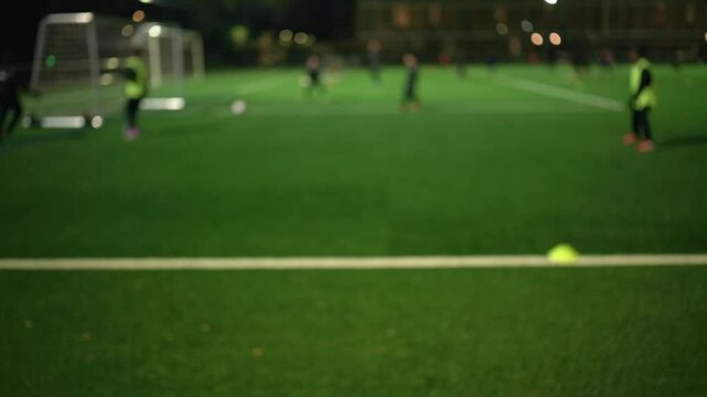 Night view of a football training session on an artificial turf field under bright floodlights with small goals and cones set up for youth practice.
