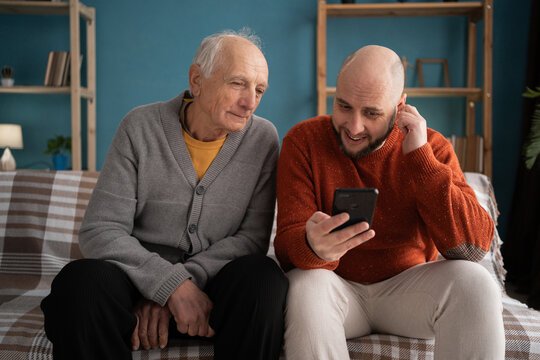 senior father with his adult son looking at a smartphone shopping online scrolling social media on sofa at home