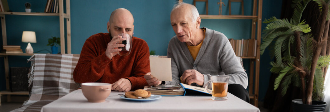 family at home. old father with adult son drinking tea sitting on sofa in living room, looking photo album - Powered by Adobe