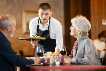Young man waiter brings order from couple elderly man and elderly woman in restaurant