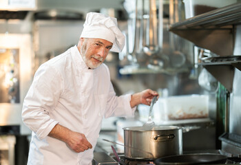 Professional senior male cook preparing dish on the stovetop in busy restaurant kitchen