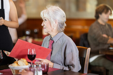 Elderly lady holds menu in hands and communicates with unrecognizable waitress in restaurants recreation area. Visitor makes order and expresses wishes.