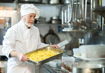 Senior man cook in kitchen is at work. Kitchen worker in white uniform performs tasks of chef and auxiliary functions, cooking French fries.