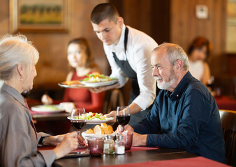 In restaurant hall, guy waiter brought order food to mature couple visitor, puts traditional Mediterrian salad of leafy greens, cheese on table. Waiter fetch and carry food for restaurant client.