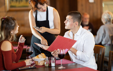 At cafe hall, Filipino girl waitress communicates with girl and boyfriend customers, take order, answer questions about composition of dish of day. Client consult with waiter about vegan health menu .