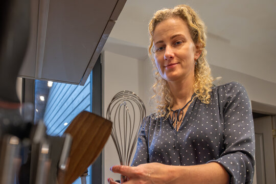 Woman with curly hair is preparing healthy food in a modern kitchen, holding a whisk and wooden spoon, showcasing cooking tools and vibrant ingredients for nutritious meal preparation