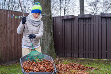 Woman with curly hair wearing a winter hat is raking autumn leaves in a backyard, surrounded by...