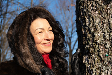 Mid-age woman in a luxurious fur coat smiles joyfully outdoors during winter, surrounded by nature, radiating beauty and warmth in a serene atmosphere
