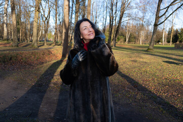 Mid-age woman in a luxurious fur coat smiles joyfully outdoors during winter, surrounded by trees and sunlight, capturing the essence of beauty and happiness in nature