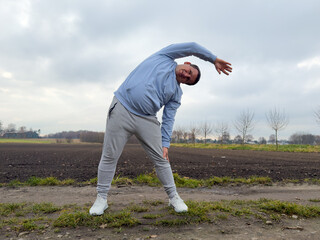 A man stretching sideways outdoors in a rural setting, wearing casual sportswear, with a cloudy sky and fields in the background