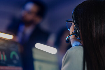 Close-up rear view of young female operator wearing headset with microphone in blue-lit room,...