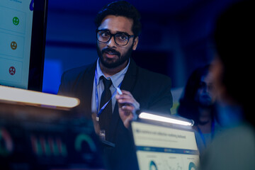 Focused Indian businessman in glasses analyzing financial data charts on night shift in blue-lit modern office, working on urgent corporate project with team.