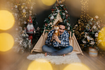 A smiling, dark-haired man with wrinkles on his face, wearing jeans and a shirt, sits near a...