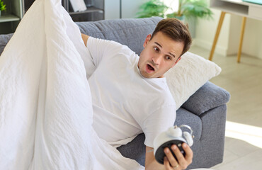 Man checks alarm clock, feeling shock at morning. He wakes in bed, realizing a wakeup gone late, oversleeping with a shocked look in a bright room. Concept: urgency, waking, time pressure.