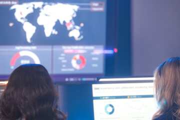Rear view of female analysts monitoring global data maps and business charts on high-tech dashboard screens. Team tracking international statistics in a modern digital command center.
