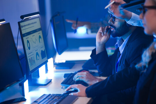 Focused Indian male operator in glasses and headset discussing data with a colleague in a blue-lit control room, managing urgent network operations or customer support. - Powered by Adobe