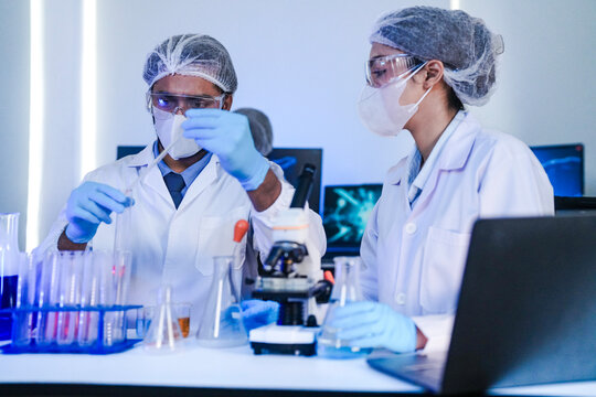 Diverse team of scientists in protective gear collaborating in modern blue lab, analyzing DNA helix structure on monitor and testing chemical samples for medical research.