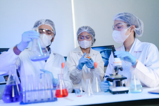 Diverse team of scientists in protective gear collaborating in modern blue lab, analyzing DNA helix structure on monitor and testing chemical samples for medical research.