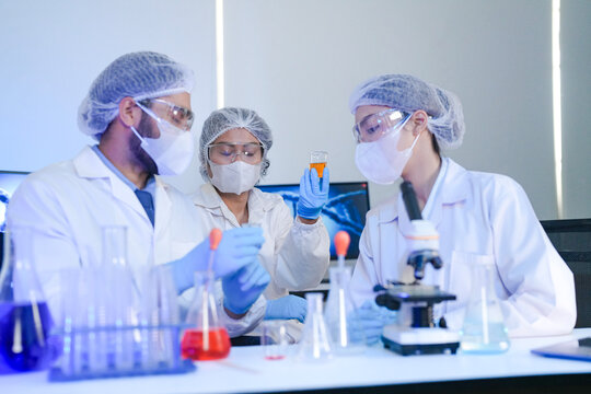 Diverse team of scientists in protective gear collaborating in modern blue lab, analyzing DNA helix structure on monitor and testing chemical samples for medical research.