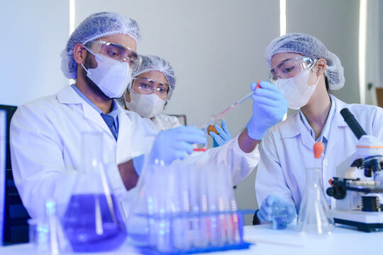 Diverse team of scientists in protective gear collaborating in modern blue lab, analyzing DNA helix structure on monitor and testing chemical samples for medical research.