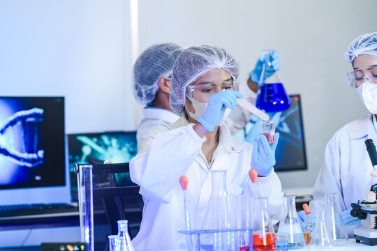 Diverse team of scientists in protective gear collaborating in modern blue lab, analyzing DNA helix structure on monitor and testing chemical samples for medical research.