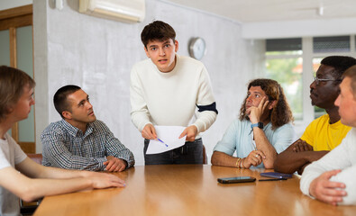 Focused young guy student holding papers ad discussing assignments or projects with interested male group of various ages and nationalities gathered around table in classroom..