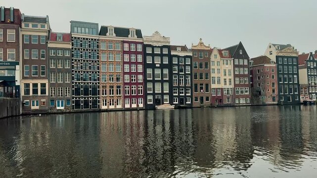 Iconic row of traditional narrow canal houses lining the water in Amsterdam, Netherlands. Historic Dutch architecture with colorful facades reflects in the calm canal on a gray overcast day.

