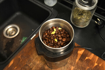 Coffee beans are placed in a grinder located on a kitchen countertop. A jar of herbs is visible in the background, emphasizing a cozy kitchen environment