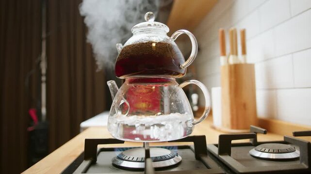 Steam rises as dark black tea brews in a transparent glass teapot placed on a lit gas stove in cozy kitchen.
